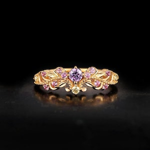 May include: A gold-colored ring with a central purple gemstone and surrounding pink and yellow gemstones. The ring features an ornate, floral design with intricate details. The ring is set against a black background.