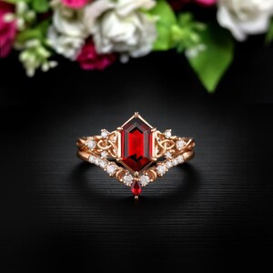 May include: A rose gold ring set with a large, rectangular, deep red gemstone. The engagement ring has a delicate band with small, clear accent stones. A matching band with a curved design completes the set. The ring set is suitable for a special occasion.