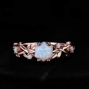 May include: Rose gold ring featuring a central, hexagonal opal stone, surrounded by smaller opals and delicate leaf-like details. The ring's band and setting have a warm, metallic hue, creating an elegant and nature-inspired design. The ring is set against a dark background.