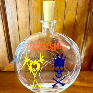 May include: A clear, round glass bottle with a wooden stopper. The bottle features the word "PEARLJAM" in orange, along with yellow and blue graphics. The graphics include a stick figure and a skull with crossbones.