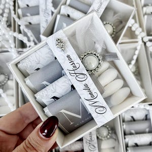 Op de afbeelding: Een witte doos met een decoratief lint en een zilveren broche, met verpakte chocolaatjes en witte snoepjes. De doos is versierd met een bloemmotief en de tekst "Manolia Hassan" en de datum "05.10.2023".
