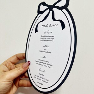 Peut inclure: Une carte de menu ovale avec une bordure noire et un motif de n&oelig;ud en haut. Le menu &eacute;num&egrave;re les entr&eacute;es, les plats, les accompagnements et les desserts, y compris des &eacute;l&eacute;ments comme le steak, le homard mac & cheese et les cupcakes. Le texte est manuscrit.