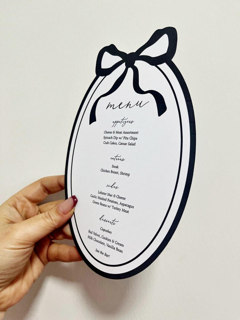 Peut inclure: Une carte de menu ovale avec une bordure noire et un motif de n&oelig;ud en haut. Le menu &eacute;num&egrave;re les entr&eacute;es, les plats, les accompagnements et les desserts, y compris des &eacute;l&eacute;ments comme le steak, le homard mac & cheese et les cupcakes. Le texte est manuscrit.