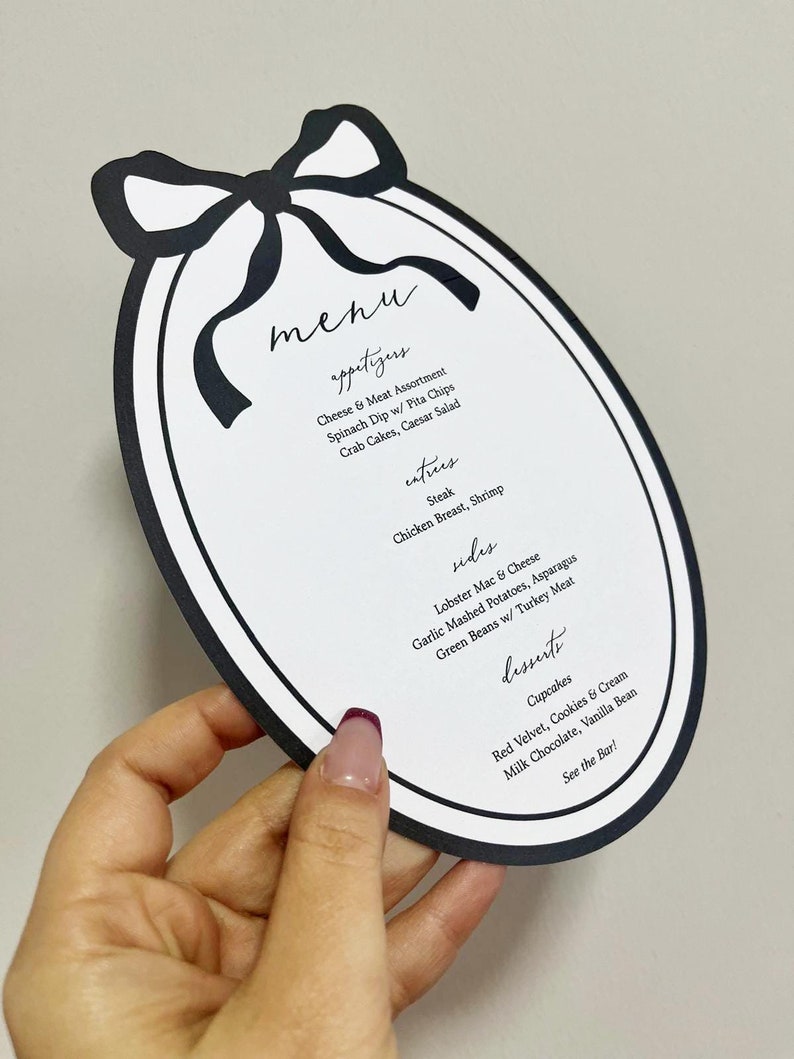Peut inclure: Menu ovale noir et blanc, surmont&eacute; d'un n&oelig;ud. La carte pr&eacute;sente des entr&eacute;es, des plats, des accompagnements et des desserts, notamment du steak, des cupcakes et du mac and cheese au homard.