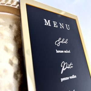 Puede incluir: Un tablero de men&uacute; negro enmarcado con un marco dorado. El men&uacute; est&aacute; escrito en blanco e incluye los siguientes art&iacute;culos: Ensalada, ensalada de la casa, Pasta, penne a la vodka, Entradas.
