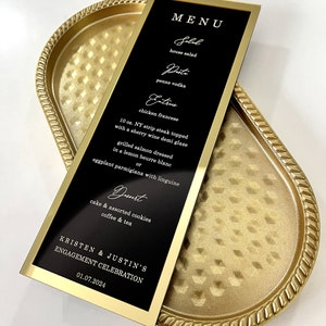 Puede incluir: Una tarjeta de men&uacute; negra y dorada con un marco dorado. El men&uacute; enumera los alimentos y bebidas para la celebraci&oacute;n de compromiso de Kristen y Justin el 01.07.2024. El men&uacute; incluye ensalada, pasta, platos principales y postre.