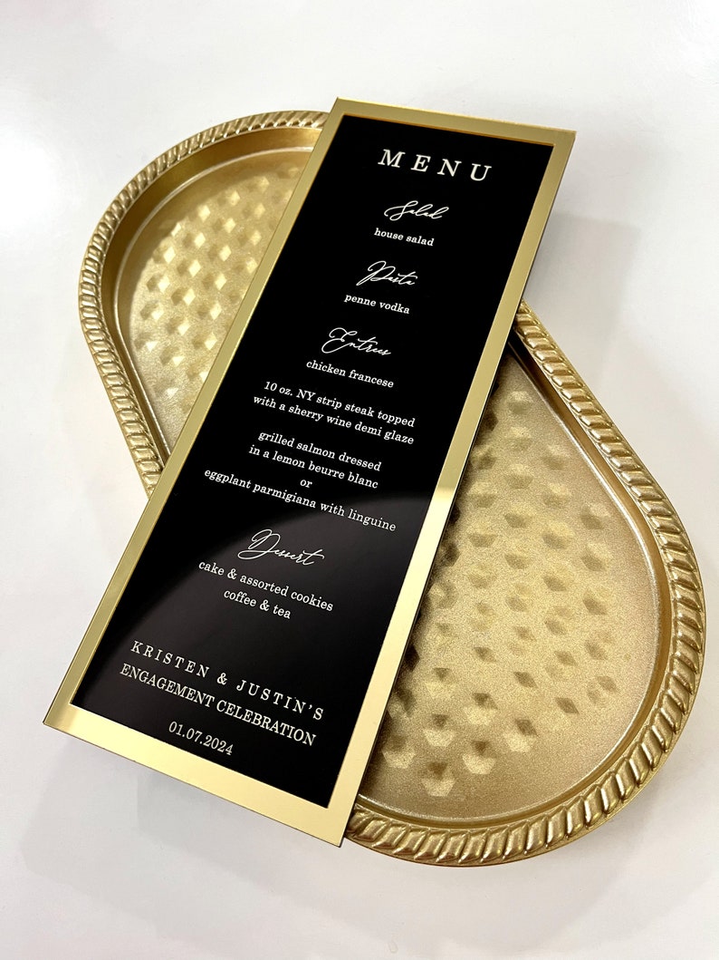 Puede incluir: Una tarjeta de men&uacute; negra y dorada con un marco dorado. El men&uacute; enumera los alimentos y bebidas para la celebraci&oacute;n de compromiso de Kristen y Justin el 01.07.2024. El men&uacute; incluye ensalada, pasta, platos principales y postre.
