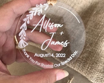 May include: A clear round acrylic ornament with a white floral design and the text "Allison + James August 14, 2022 Thank You For Sharing Our Special Day."