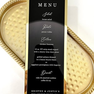 Puede incluir: Un men&uacute; de boda negro con marco dorado y texto blanco, que enumera el men&uacute; para la celebraci&oacute;n de compromiso de Kristen y Justin. El men&uacute; incluye opciones de ensalada, pasta, platos principales y postres. La fecha del evento es el 01.07.2024.
