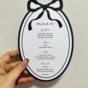 Peut inclure: Un menu en forme ovale avec un n&oelig;ud noir en haut. Le menu comprend des entr&eacute;es, des plats principaux, des accompagnements et des desserts. Il y a du steak, du poulet et des cupcakes.