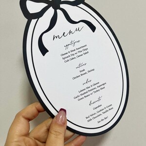 Peut inclure: Menu ovale noir et blanc avec un motif de n&oelig;ud en haut. La carte propose des entr&eacute;es, des plats principaux, des accompagnements et des desserts. Le menu comprend des plats comme le steak et les cupcakes.