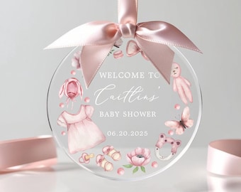 May include: A pink and white baby shower ornament with the text "Welcome to Caitlin's Baby Shower 06.20.2025". The ornament features a variety of baby-themed illustrations, including a dress, a hat, a rattle, a butterfly, and a teddy bear.
