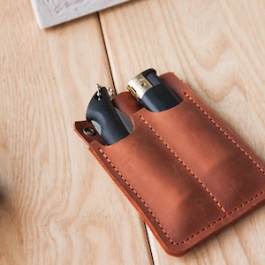 May include: A brown leather pocket organiser with two slots for carrying a pocket knife and a lighter. The organiser is made of brown leather and has a stitched edge.