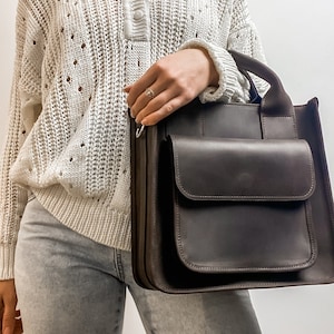 May include: A brown leather tote bag with a large front pocket and a top handle. The bag is being held by a person wearing a white sweater and light blue jeans.