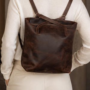 May include: A dark brown leather backpack with a smooth, matte finish and two shoulder straps. The bag features a rectangular shape with rounded corners and visible stitching along the top edges where the straps are securely attached to the body.