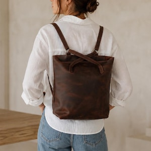 Puede incluir: Una mochila de cuero marrón oscuro con forma rectangular vertical y acabado liso. El bolso cuenta con dos correas ajustables para los hombros que se cruzan en la parte superior, unidas por anillos metálicos, y un compartimento principal sencillo.