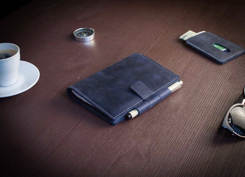 Op de afbeelding: Een donkerblauw leren notitieboekje met een pennenhouder en een klep sluiting. Het notitieboekje is gesloten en ligt op een bruin houten oppervlak.