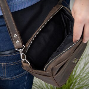 May include: A brown leather crossbody bag with a top zipper closure, a black fabric interior lining, and a matching adjustable leather shoulder strap attached with silver-toned metal hardware. The bag features a small rectangular metal accent plate.