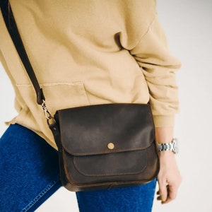 K&ouml;nnte beinhalten: Eine braune Leder-Umh&auml;ngetasche mit Klappenverschluss und einem einzigen Riemen. Die Tasche hat eine kleine Tasche auf der Vorderseite.