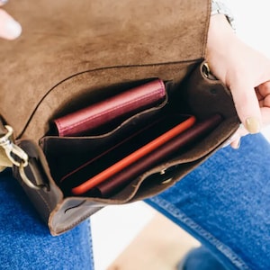 Pu&ograve; includere: Una borsa a tracolla in pelle marrone con chiusura a lembo e tracolla. La borsa &egrave; aperta e ha tre scomparti, di cui due sono pieni di portafogli rossi e rosa e un telefono.