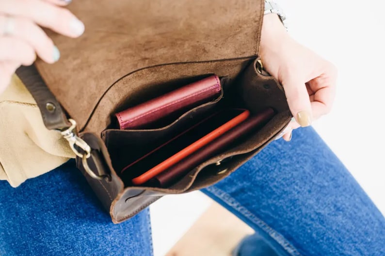 Pu&ograve; includere: Una borsa a tracolla in pelle marrone con chiusura a lembo e tracolla. La borsa &egrave; aperta e ha tre scomparti, di cui due sono pieni di portafogli rossi e rosa e un telefono.