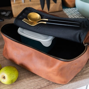 May include: A brown leather zippered pouch with a black fabric lining. The pouch is open and contains a white plastic container, a gold spoon, and a gold fork with black handles.