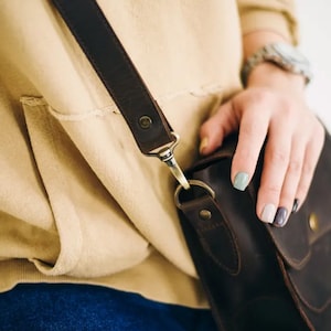 Pu&ograve; includere: Una borsa a tracolla in pelle marrone con una lunga tracolla e una chiusura a lembo. La borsa ha una piccola tasca sul davanti e viene indossata sulla spalla.
