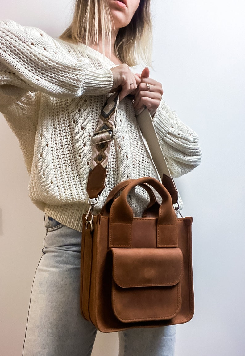 May include: A brown suede tote bag with a top handle and a long adjustable strap. The strap is patterned with brown, pink and white geometric shapes. The bag has a large front pocket.