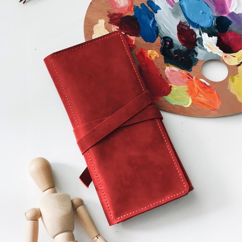 May include: A red leather wallet with a strap closure. The wallet is closed and lying on a white surface.