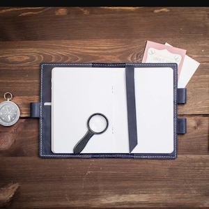 Op de afbeelding: Een blauw lederen gebonden dagboek met een blanco pagina, een vergrootglas, een kompas, een pen en twee papieren met de tekst "About You" erop.