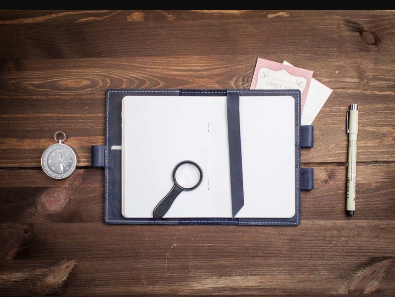 Op de afbeelding: Een blauw lederen gebonden dagboek met een blanco pagina, een vergrootglas, een kompas, een pen en twee papieren met de tekst "About You" erop.