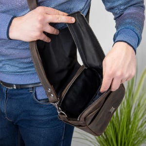 May include: A dark brown leather crossbody bag with a vertical zippered opening, revealing a black fabric interior lining. The bag features a rectangular metal logo plate on the front exterior and a sturdy adjustable strap attached with metal hardware.