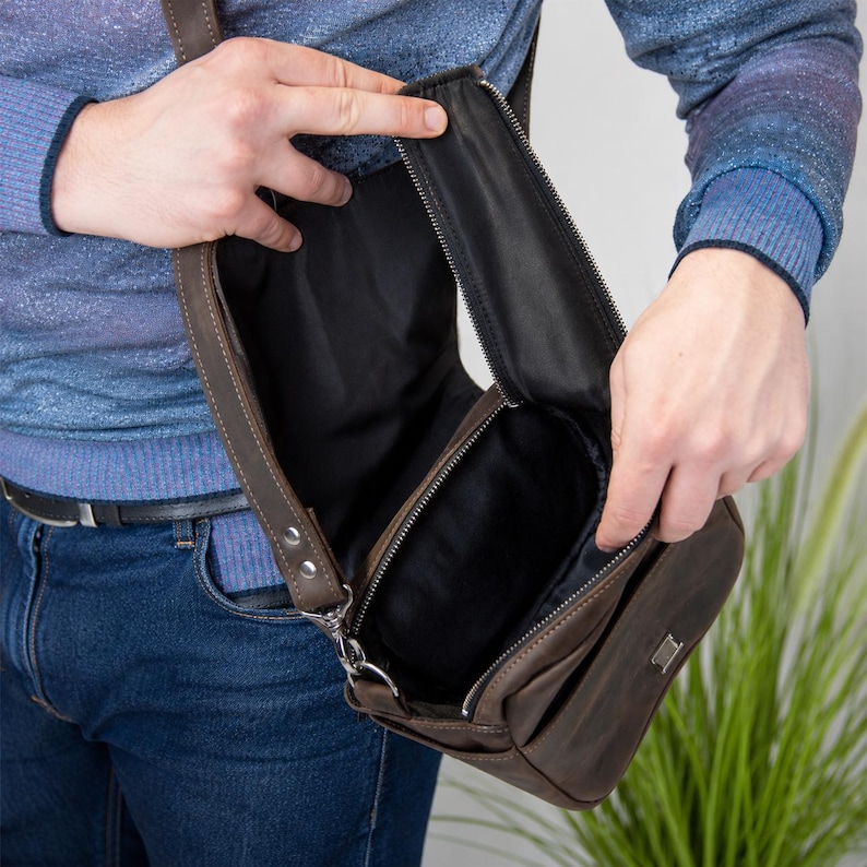 May include: A dark brown leather crossbody bag with a vertical zippered opening, revealing a black fabric interior lining. The bag features a rectangular metal logo plate on the front exterior and a sturdy adjustable strap attached with metal hardware.
