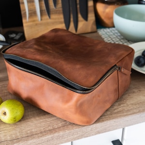 May include: A brown leather zippered pouch with a black zipper. The pouch is open and the inside is lined with black fabric.