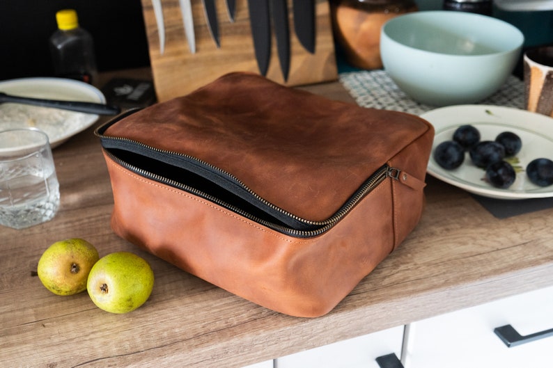 May include: A brown leather zippered pouch with a black zipper. The pouch is open and the inside is lined with black fabric.