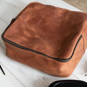 May include: A brown leather zippered pouch or bag. The pouch is on a white wooden surface with a gold fork and spoon and a green pear.