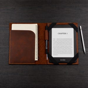 K&ouml;nnte beinhalten: Eine braune Lederh&uuml;lle mit einem spiralgebundenen Notizbuch auf der linken Seite und einem E-Reader auf der rechten Seite. Der Bildschirm des E-Readers zeigt den Text CHAPTER 1 und einen Blindtext. Ein schwarzer Stift steckt in einer Schlaufe, ein silberner Stift liegt daneben.