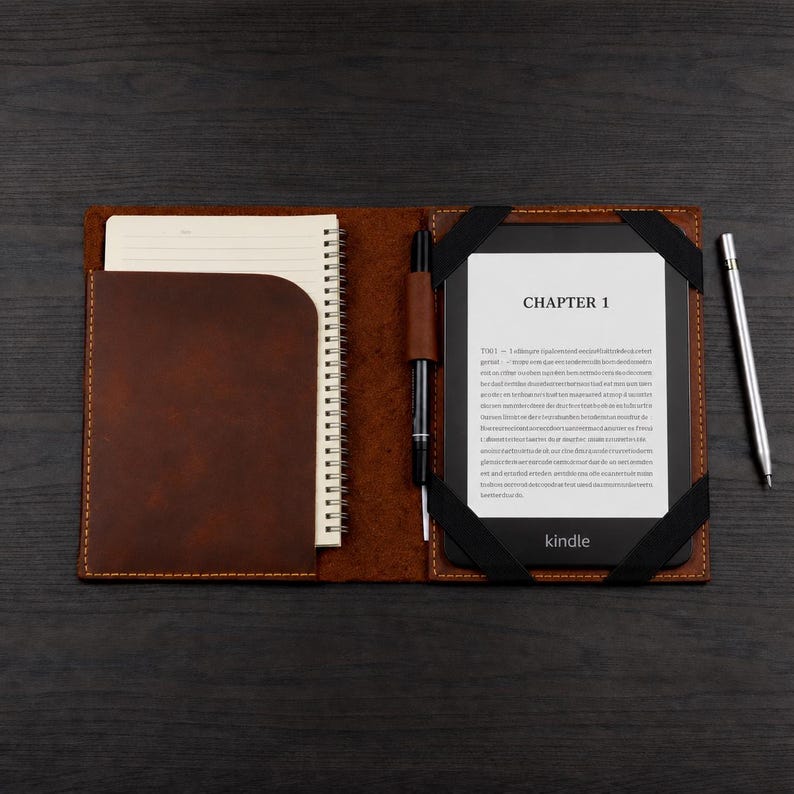 K&ouml;nnte beinhalten: Eine braune Lederh&uuml;lle mit einem spiralgebundenen Notizbuch auf der linken Seite und einem E-Reader auf der rechten Seite. Der Bildschirm des E-Readers zeigt den Text CHAPTER 1 und einen Blindtext. Ein schwarzer Stift steckt in einer Schlaufe, ein silberner Stift liegt daneben.