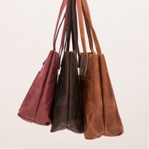 Puede incluir: Tres bolsos tote de cuero en diferentes tonos, incluyendo burdeos, marr&oacute;n oscuro y tostado. Cada bolso cuenta con correas largas para el hombro, una base rectangular y un bolsillo exterior visible en el lateral, colgados juntos sobre un fondo liso.