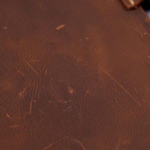 K&ouml;nnte beinhalten: Eine Nahaufnahme einer braunen Lederoberfl&auml;che mit einer nat&uuml;rlichen, matten Textur und sichtbaren hellen Kratzern und Schrammen auf der Narbung, die ein abgenutztes Aussehen mit Variationen in den dunkelbraunen Farbt&ouml;nen aufweist.