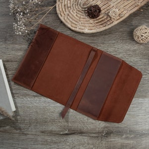 May include: A brown leather book cover with a strap closure. The cover is open to reveal the inside, which is lined with a brown fabric.