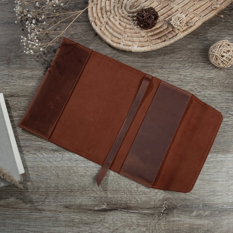 May include: A brown leather book cover with a strap closure. The cover is open to reveal the inside, which is lined with a brown fabric.