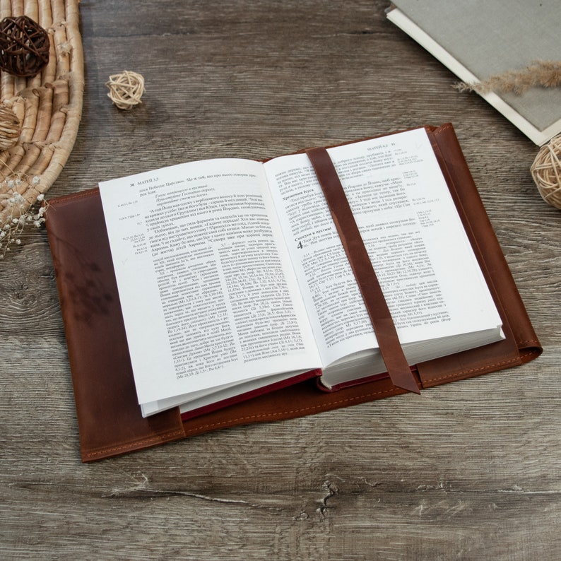 May include: A brown leather-bound book with a red bookmark. The book is open to a page with text in Russian. The book is lying on a wooden surface.