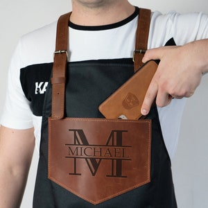 May include: A black leather apron with a brown leather pocket. The pocket has a large "M" with the name "MICHAEL" underneath it. A phone is in the pocket.