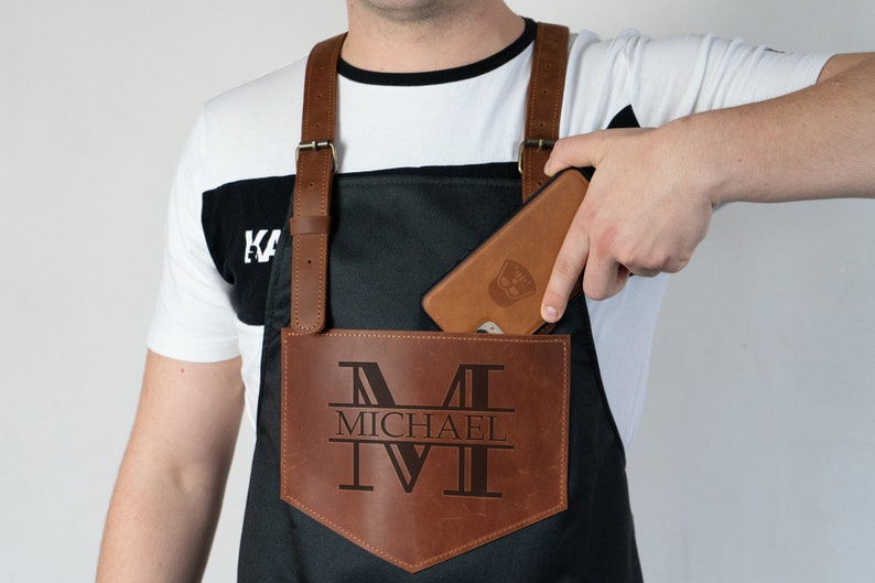 May include: A black leather apron with a brown leather pocket. The pocket has a large "M" with the name "MICHAEL" underneath it. A phone is in the pocket.