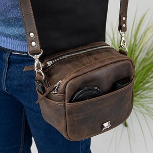 May include: A dark brown leather crossbody bag with a rectangular shape, featuring a top flap, silver-toned metal hardware, a zippered main compartment, and an open front pocket containing a black circular lens cap and a small black rectangular object.