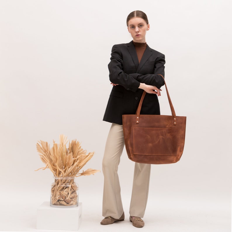 Puede incluir: Un bolso tote grande y rectangular de cuero marr&oacute;n con dos correas largas para el hombro, un compartimento principal espacioso y un bolsillo frontal exterior. El cuero tiene un acabado mate suave con grano natural y variaciones tonales.