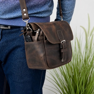 May include: A dark brown leather crossbody bag featuring a fold-over flap with a metal buckle closure, an adjustable shoulder strap, and an exterior side pocket containing a small black electronic device with a visible button.