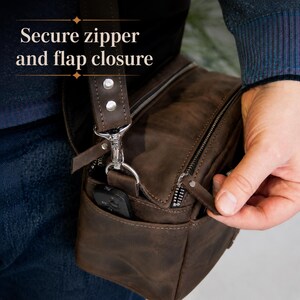 May include: A brown leather shoulder bag featuring a secure zipper and flap closure. The bag includes a silver metal swivel clip attaching the shoulder strap to the main body, with a small side pocket containing a black electronic device.