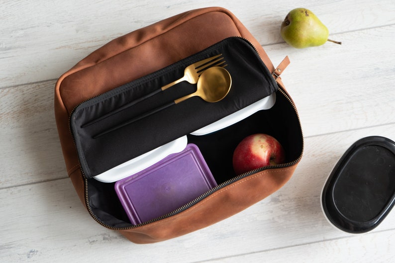May include: A brown leather lunch bag with a zipper closure. The bag is open and contains a black utensil holder with a gold fork and spoon, two white containers, a purple container, and a red apple.